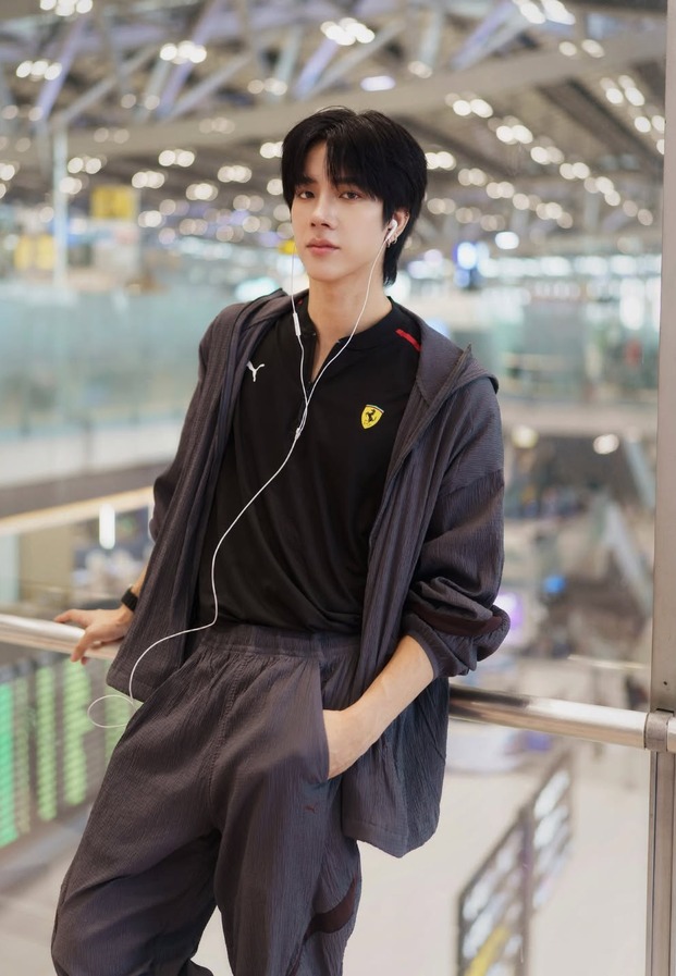 แฟรงค์สวมเสื้อโปโลสีดำทับด้วยแจ็กเก็ตซิปสีเทายืนในอาคารสนามบิน